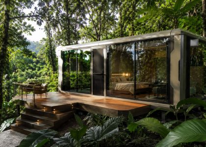A white modular cabin with floor-to-ceiling glass doors and a large wooden deck built directly into a dense rainforest setting.